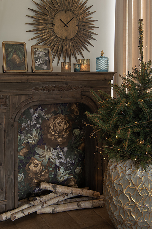 Un manteau de cheminée marron avec deux cadres photo dorés, trois porte-bougies et un bocal en verre bleu, et devant lui, un petit sapin de Noël dans un pot en métal