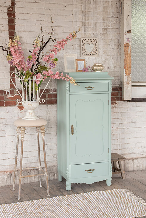 Un meuble de rangement vert menthe avec trois cadres photo campagnards, un porte-bougie chauffe-plat, et à côté se trouve un tabouret shabby chic et un vase blanc en fer forgé avec des fleurs artificielles