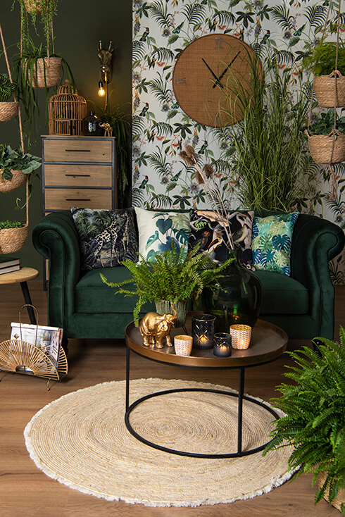Un canapé vert avec quatre coussins décoratifs, et devant se trouve une table basse dorée remplie de photophores, de pots de fleurs et de figurines, posée sur un tapis rond