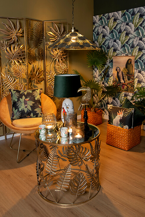 Une table basse thématique botanique avec des photophores et une lampe de table, et à côté se trouve une chaise jaune avec un coussin décoratif