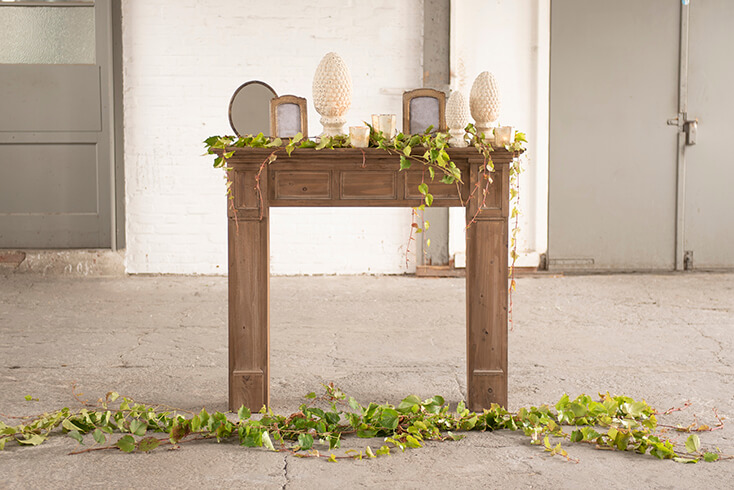 A wooden fireplace mantel with decorative sculptures, photo frames, candle holders, and a table mirror, adorned with artificial greenery