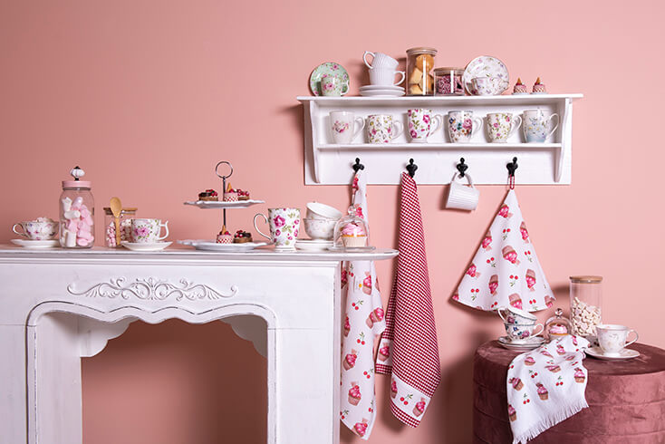 A white fireplace mantel with tableware, cake stands, and pastries, and behind the mantel, there's a white wall cabinet with mugs, tableware, and kitchen textiles