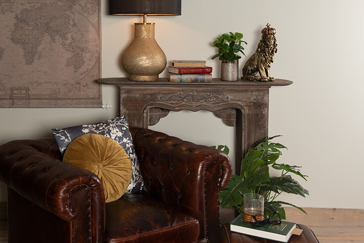 A brown fireplace mantel with a large table lamp, books, a flowerpot, and a lion sculpture. In front of the mantel, there's a leather armchair with two decorative cushions