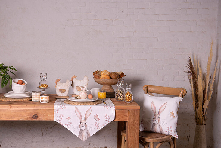 A set Easter table with tableware, storage jars, a table runner, and a cake platter with pastries, and on the dining chair, there is a decorative cushion