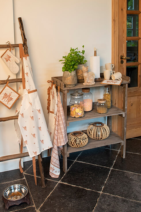 A console table filled with storage jars, lanterns, and kitchen accessories, and next to the console table, there is a wooden ladder with kitchen textiles hanging on it