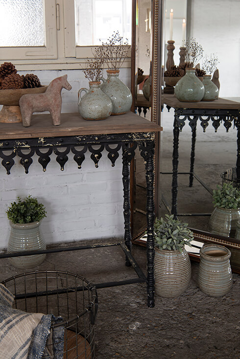 A rustic console table with vases, a horse sculpture, and a cake platter, and next to it, there is a full-length mirror with flower pots on the floor