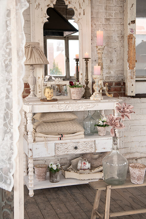 Un style d'intérieur shabby chic avec une console blanche, des accessoires décoratifs pour la maison, une lampe de table, des coussins et des bougeoirs