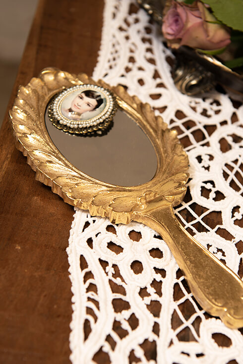 A gold-colored hand mirror with a brooch featuring Audrey Hepburn