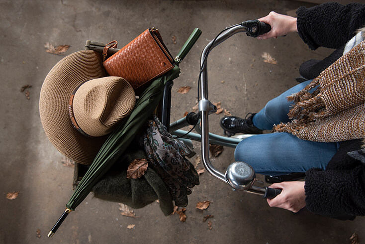 Iemand op een fiets met in het mandje een bruine hoed, groene paraplu, herfstsjaal en een bruine handtas