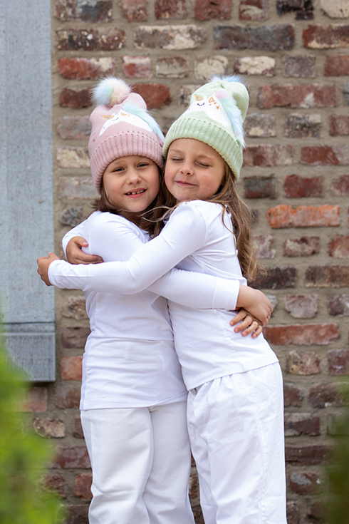 Twee meiden die elkaar knuffelen en een kindermuts dragen met een eenhoorn in het roze en groen