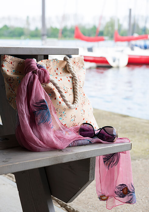 Un tavolo da picnic con una borsa da spiaggia beige, occhiali da sole e una sottile sciarpa estiva rosa