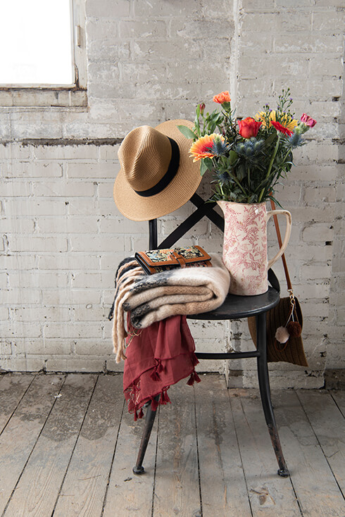 Een zwarte stoel met daarop een beige hoed, een portemonnee, en dikke wintersjaal en een schenkkan met een bos bloemen