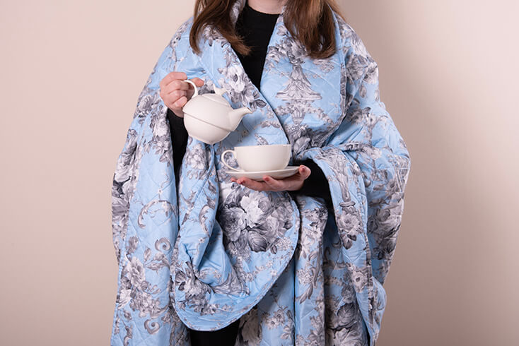 Someone wearing a blue bedspread with black and white flowers and holding a tea for one teapot