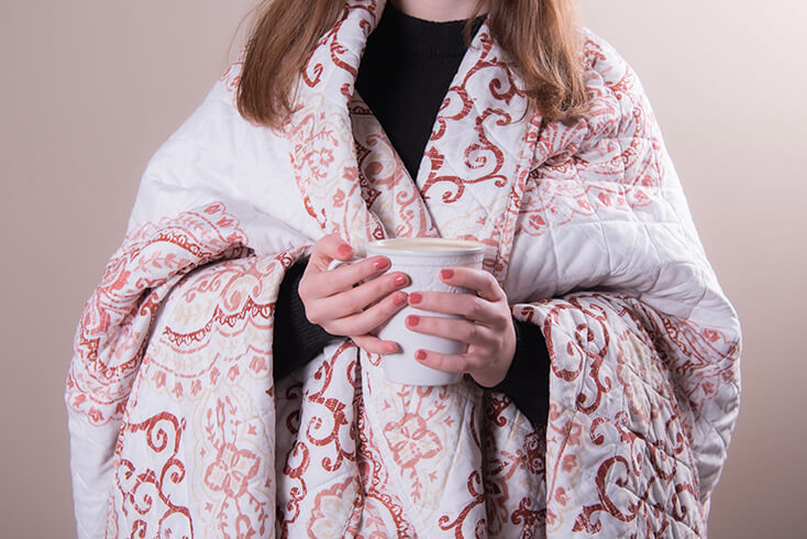 A folded shabby chic bedspread, and the person is holding a rural mug with coffee