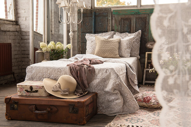 Een shabby chic slaapkamer met een opgemaakt bed van bedspreien en veel kussens en voor het bed staat een antieke koffer met een zomerhoed