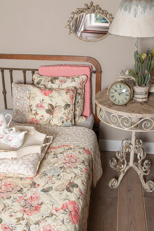 Een landelijke slaapkamer met een vintage bedsprei en slaapkamer kussens en naast het bed staat een shabby chic nachtkastje met een wekker