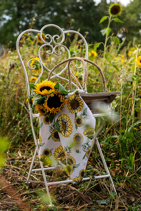 Ein weißer Gartenstuhl mit einem Holzkorb darauf, der einen Strauß Sonnenblumen und ein Sonnenblumen-Geschirrtuch enthält