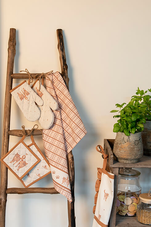 A wooden ladder with kitchen textiles, including a cat-themed tea towel, a children's oven mitt, oven gloves with cats, and children's pot holders with cats