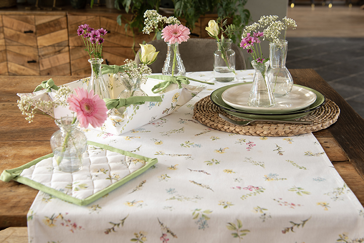 A set table with floral textiles, a flower table runner, a flower breadbasket, all accompanied by glass vases filled with pink flowers
