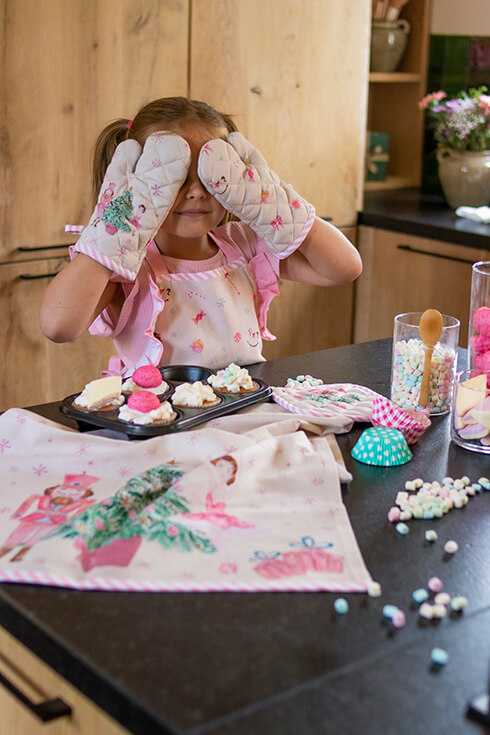 A girl in the kitchen baking with Christmas-themed textiles, wearing a children's apron and two oven mitts with a Christmas motif