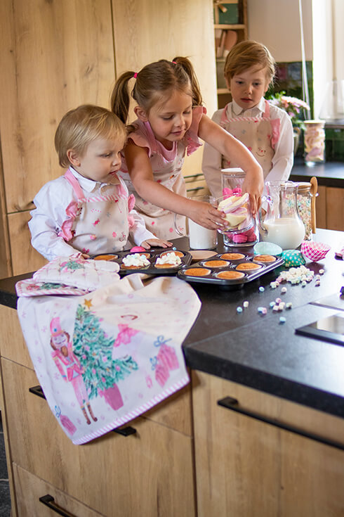Drei Kinder in der Küche, die mit Kinderschürzen und einem rosa Weihnachts-Geschirrtuch backen