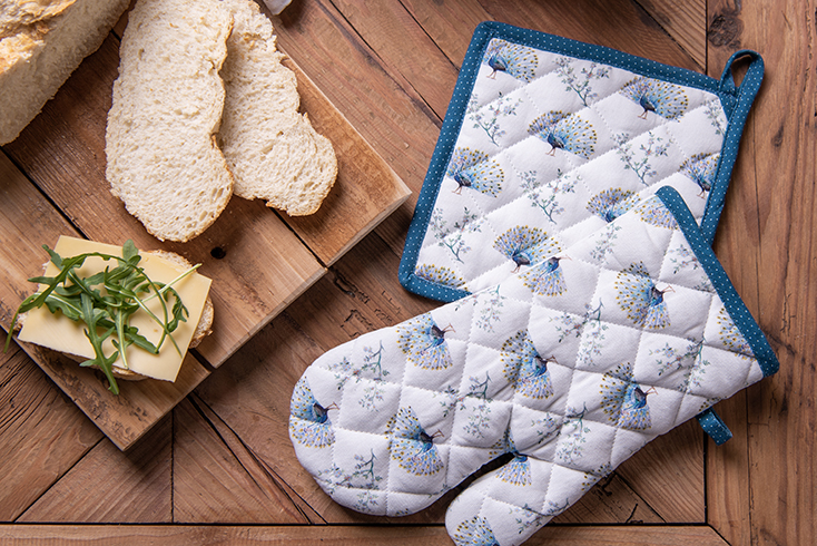 A wooden cutting board with sliced bread and a peacock-themed pot holder and peacock-themed oven mitt