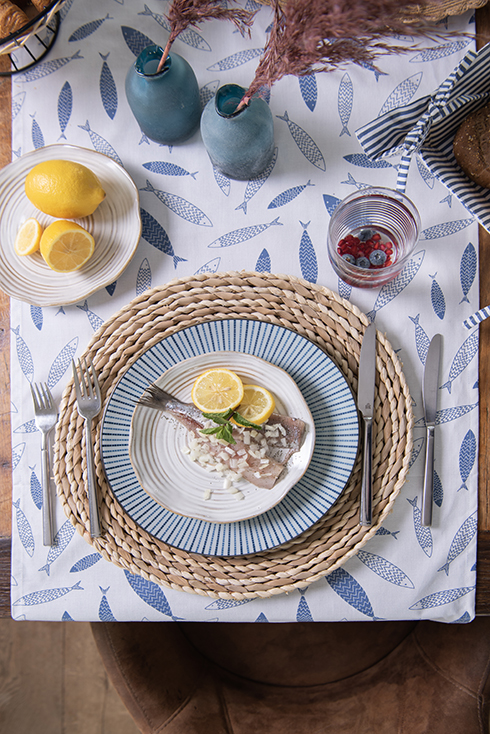 Een maritiem gedekte tafel met een ontbijtbord en dinerbord en een ronde rieten placemat en daaronder een tafelloper met visjes