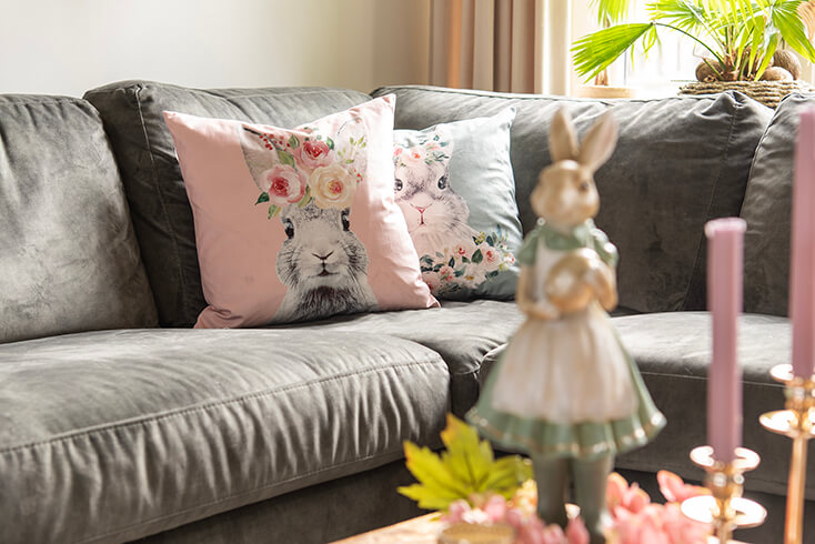 Un divano grigio con un cuscino pasquale rosa con conigli e un cuscino pasquale verde con un coniglio pasquale