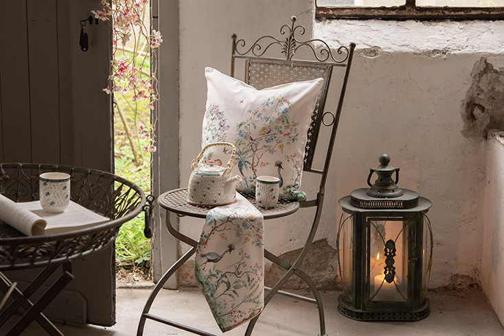 Een romantische tuin met een ijzeren tuinstoel met daarop een romantische sierkussen en theedoek en achter staat een romantische lantaarn