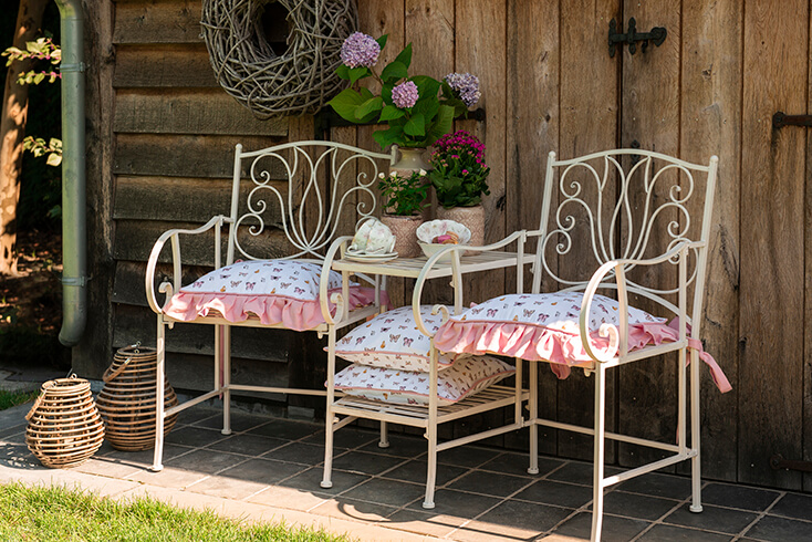 Zwei Gartenstühle mit einem kleinen Tisch draußen im Garten mit Blumentöpfen, Vasen und Geschirr. Die Stühle haben Stuhlkissen mit Schmetterlingen darauf