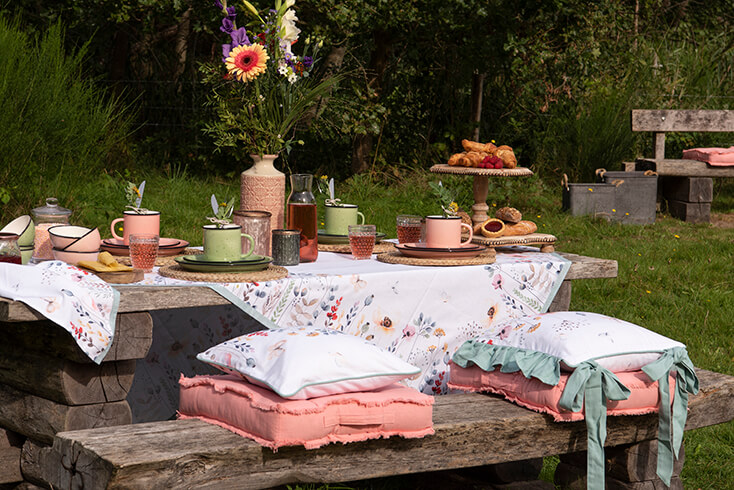 Ein Picknicktisch mit Geschirr, Küchentextilien und Küchenaccessoires. Auf der Bank liegen dicke rosa Stuhlkissen und noch weiße Stuhlkissen mit einem Blumenfeld darauf