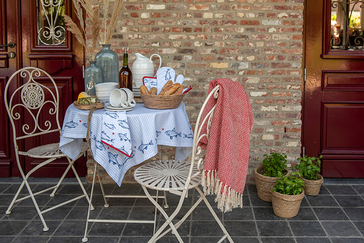 Ein Bistro-Set im Garten mit Geschirr, Küchenaccessoires, einem Tischtuch, Küchentuch und einer roten Decke, die am Gartenstuhl hängt