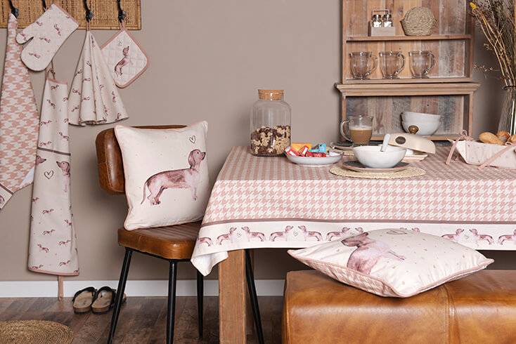 Ein modern gedeckter Esstisch mit einem Dackel-Tischtuch und Dackel-Zierkissen auf einem Ledersessel und einer Bank