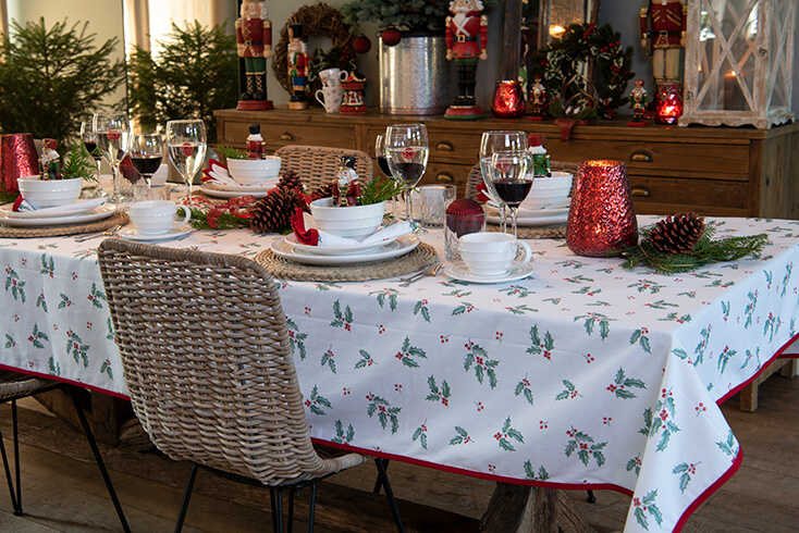 A set Christmas table with a wicker dining chair and a Christmas tablecloth with holly leaves and modern dinnerware