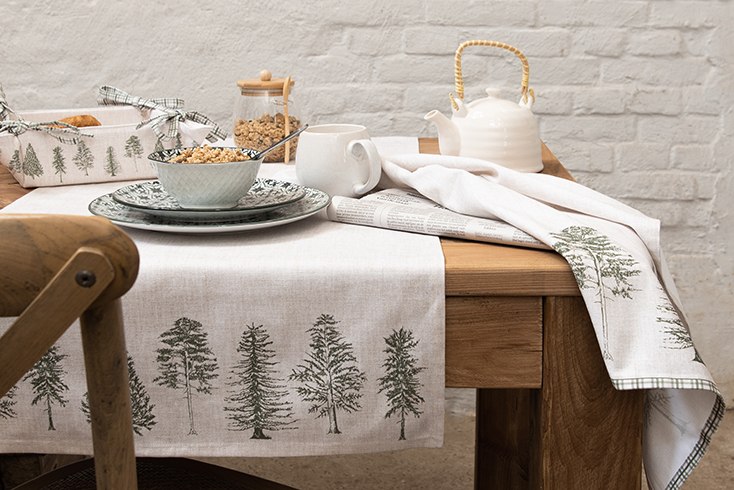 A rurally set table with a linen table runner with fir trees, dinnerware, and a linen kitchen towel with green trees