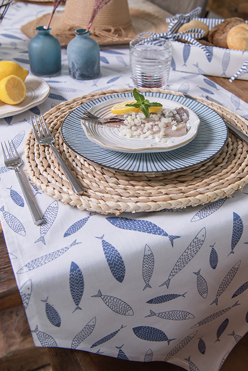 Une table dressée de style marin avec une assiette de petit-déjeuner, une assiette de dîner et une sous-assiette ronde en osier avec un chemin de table représentant des poissons