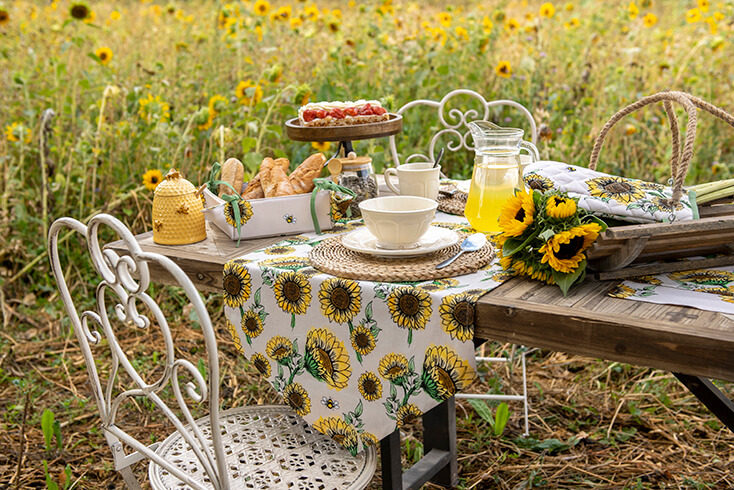 Ein gedeckter Tisch im Garten mit einem Sonnenblumen-Thema; ein Sonnenblumen-Tischläufer, ein Sonnenblumen-Brotkorb und ein Ofenhandschuh
