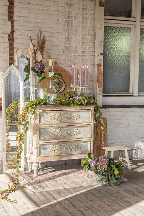 Ein Vintage-Interieurstil mit einer Kommode und Wohnaccessoires, und auf dem Boden liegt ein rechteckiger Teppich mit einem Eisengestell voller Hortensien und einem hölzernen Pflanzenständer