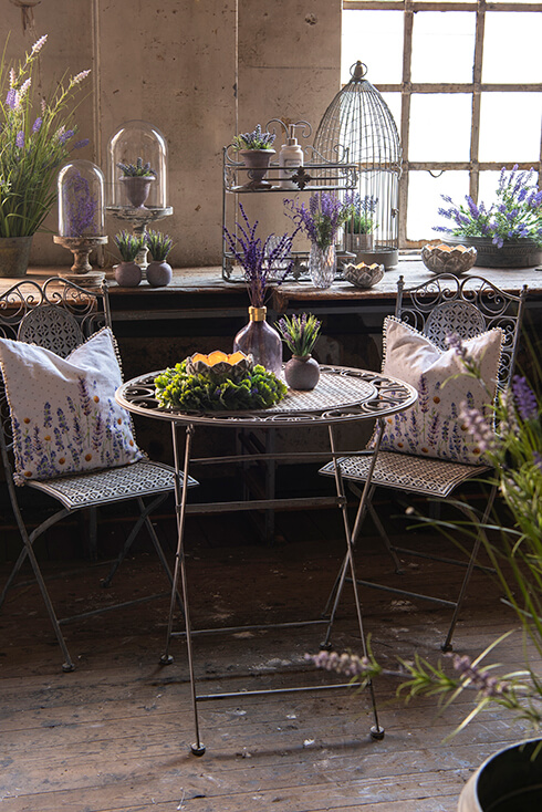 Een ijzeren bistro tafel en twee bistro stoelen met lavendel sierkussens en allerlei tuindecoratie