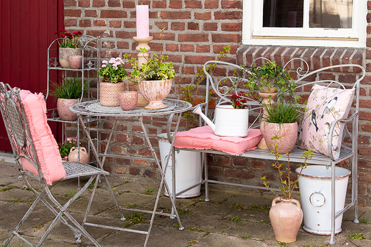 An iron garden bench with garden accessories and decorations, next to which there is an iron bistro set with pink flowerpots on the table