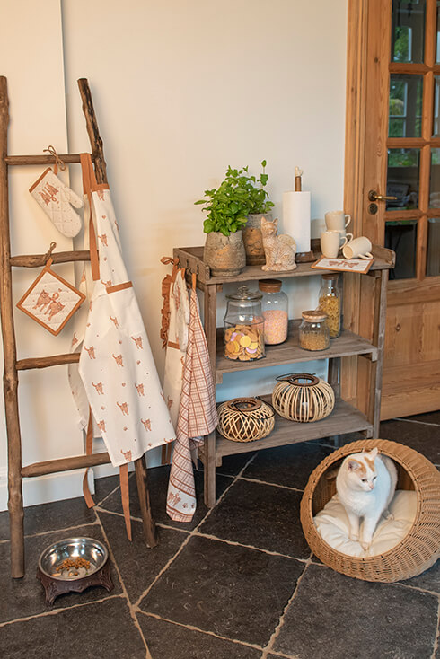 Een landelijke keuken met een houten wandtafel waar keukentextiel, voorraadpotten, rotan windlichten en keukenaccessoires op staan en een kat die in een kattenmand ligt