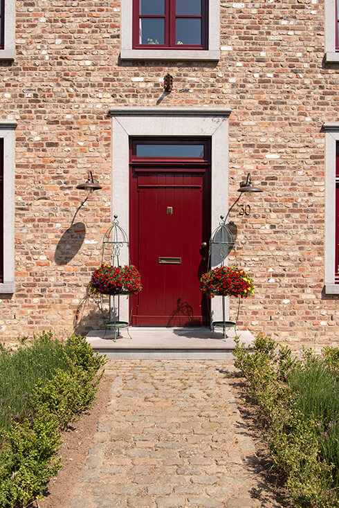 Une porte d'entrée rouge où se trouvent deux grands supports de plante en fer avec des violettes
