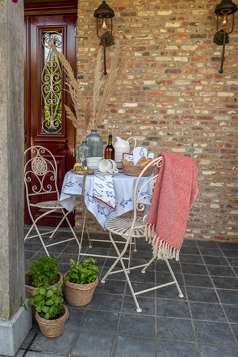 Ein Hinterhof, in dem ein romantisch gedecktes Bistro-Set mit Fischtextilien steht und eine rote Decke über dem Gartensessel hängt