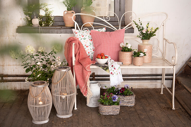 Ein romantischer Garten mit einer Schmetterling-Gartenbank und Zierkissen mit Erdbeeren, einer roten Decke, rosa Blumentöpfen, hölzernen Windlichtern und Körben mit Blumen