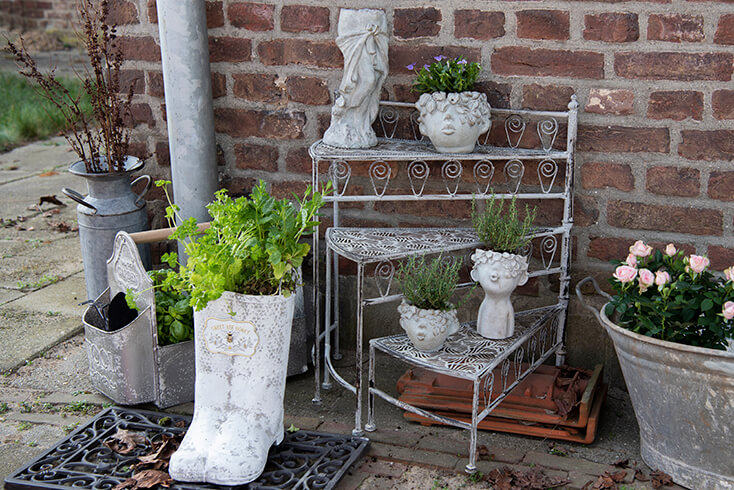 An outdoor iron rack with stone flower pots on it, along with iron boots with plants in front