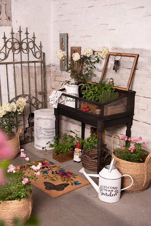 Een romantische tuin met een zwarte vitrinekast en allerlei tuindecoratie, waaronder gieters, deurmatten, bloempotten en poorten