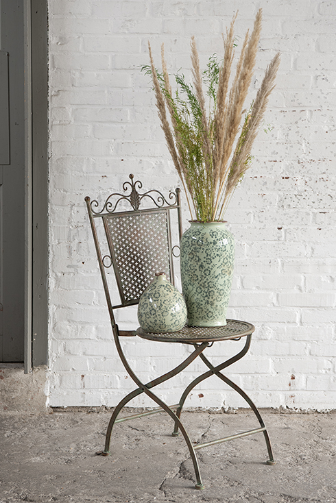 A grey bistro chair with two green vases containing a bunch of dried flowers