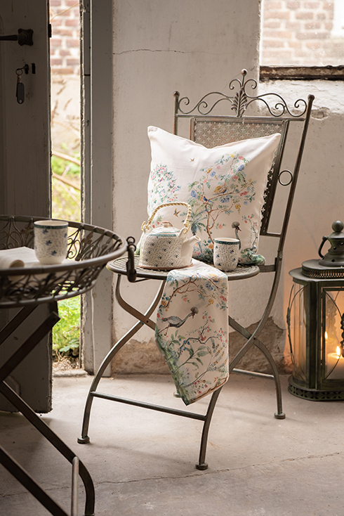 A romantic shed containing a bistro chair with decorative cushions, a tea towel, a teapot, and a coffee mug