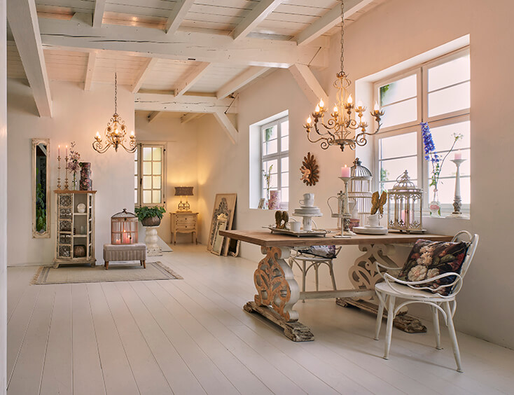 A shabby chic interior with chandeliers, a dining table, dining chairs, home decor, and a rural birdcage