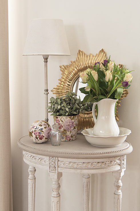 A romantic setting with a white vase, gold-framed wall mirror, decorative accessories, and a romantic table lamp with a white lampshade
